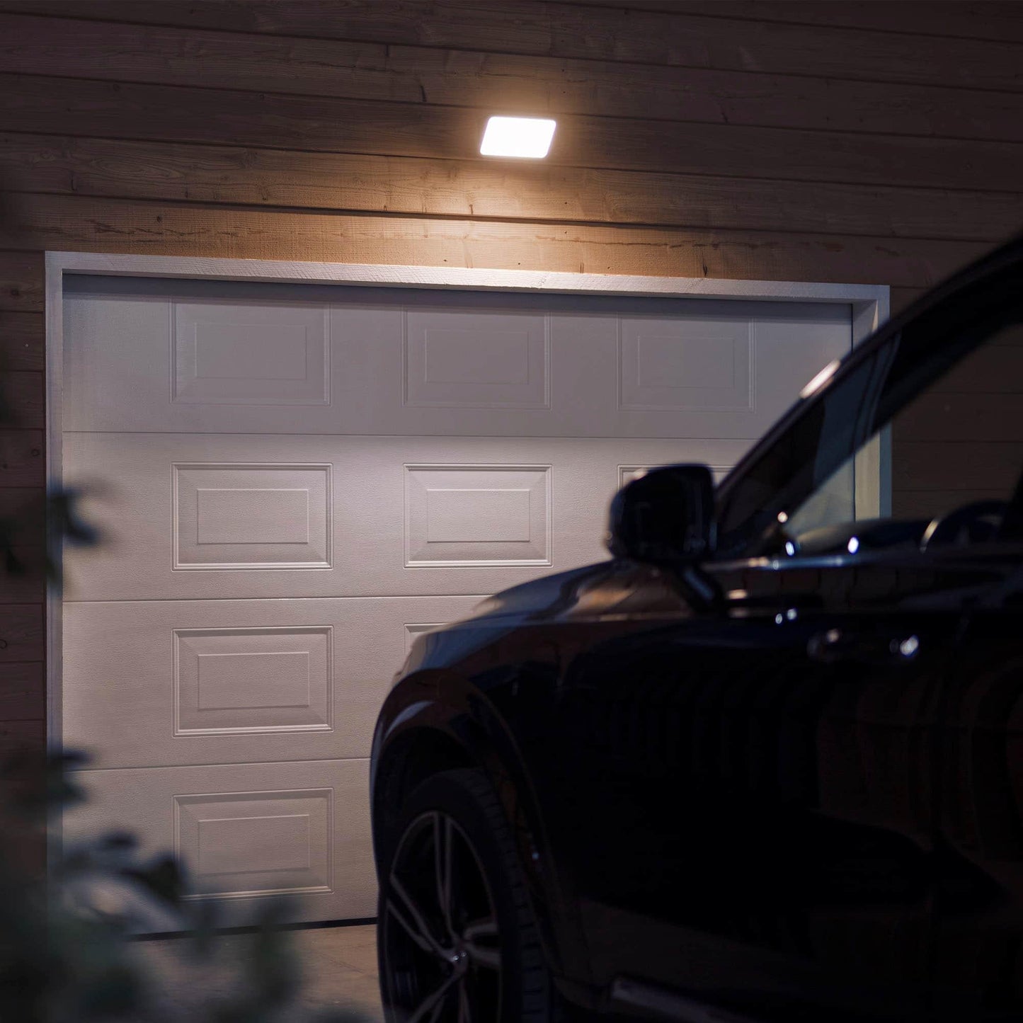 Car parked in front of a white garage door at night with a light fixture above.
